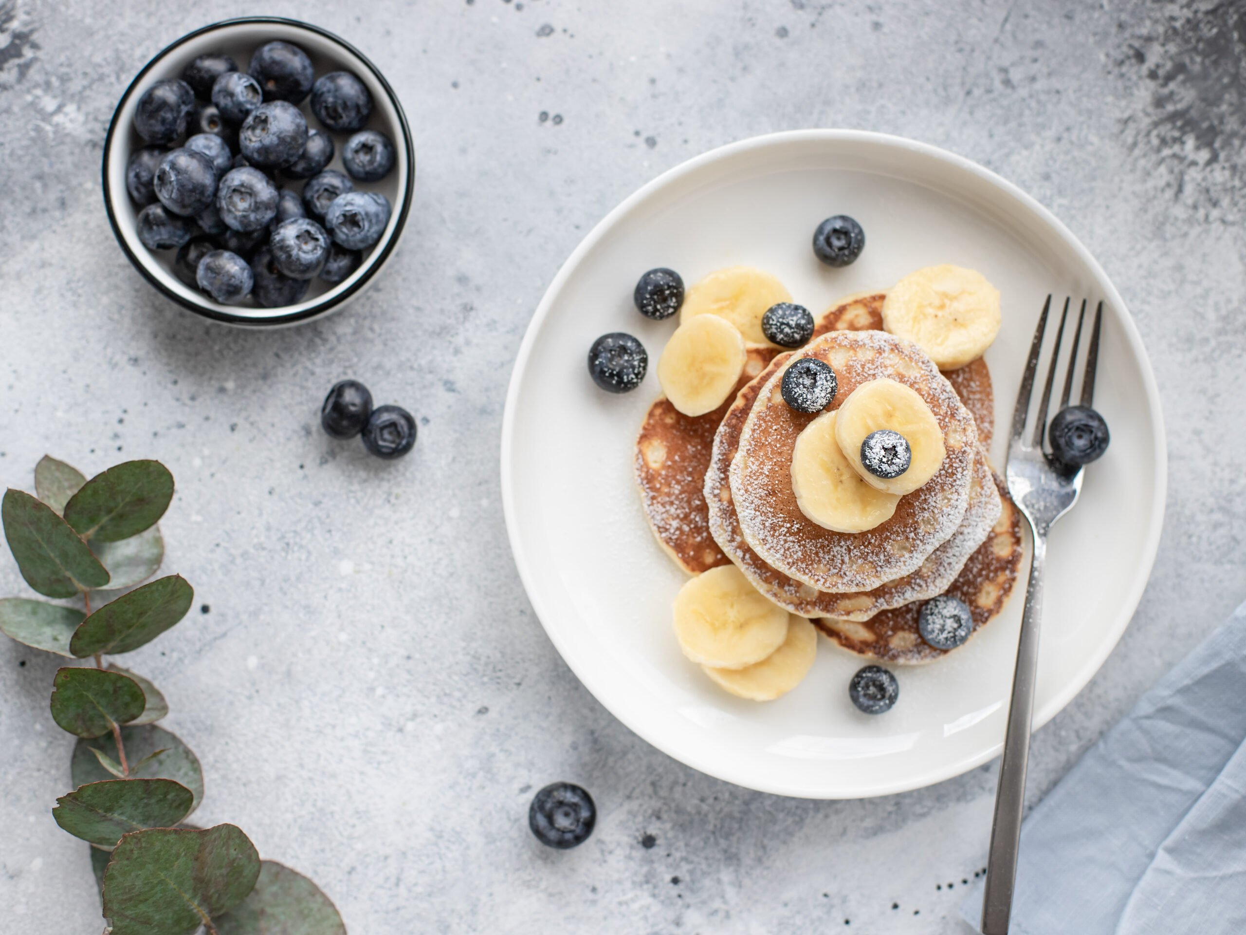 pancakes-with-fresh-blueberries-banana-in-a-white-plate-on-a-gray-background-with-a-branch-of-eucalyptus-healthy-food-concept-horizontal-image-top-view-flat-lay