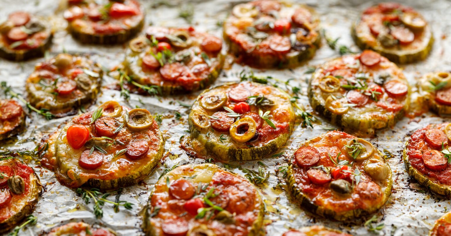 grilled-zucchini-slices-with-addition-of-mozzarella-cheese-sausage-green-olives-capers-and-herbs-on-white-baking-paper-close-up-delicious-appetizer-zucchini-pizza-bites